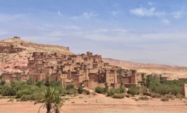14 Days Morocco Desert Tour FROM MARRAKECH ,A group of travelers on a camel trek at sunset, riding over the golden sand dunes of the Merzouga Sahara desert on a 14-day Morocco tour. The sky is ablaze with orange and pink hues, with the silhouettes of the camels and their guides creating a majestic scene. This image captures the unforgettable experience of a multi-day desert tour from Marrakech.Guests enjoying a traditional dinner under a starry sky at a luxury Berber desert camp in the Merzouga Sahara, part of an all-inclusive 14-day Morocco desert tour.Scenic drive through the High Atlas Mountains on a 14-day tour from Marrakech to the desert, showing rugged peaks and traditional Berber villages.Travelers exploring the ancient, UNESCO-listed Ait Ben Haddou kasbah, a key cultural stop on a 14-day Morocco desert tour itinerary.,A vibrant and colorful scene inside the bustling souks of Marrakech, the starting point for a comprehensive 14-day Morocco tour.A peaceful courtyard with a fountain inside a traditional Moroccan riad, showcasing typical accommodation on a 14-day cultural and desert tour.,A local guide explaining the history of the ancient medina of Fes to tourists on a 14-day imperial cities and desert tour of Morocco.